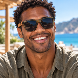 A smiling man wearing SOEK sunglasses with dark lenses and a green shirt, photographed outdoors with a beach and mountains in the background.